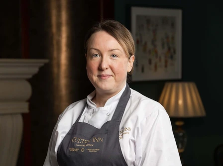 Friendly hotel staff member in restaurant setting, wearing uniform, with table set for dining.