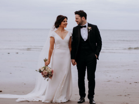 A couple in wedding attire holding hands on a serene beach near Hastings Hotels.