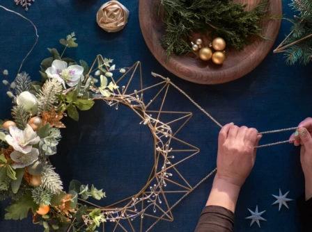 Hands crafting a festive holiday wreath with gold accents and greenery on a table.