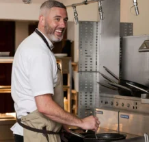 Friendly hotel chef cooking in a modern kitchen, smiling warmly at guests.