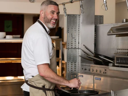 Friendly hotel chef cooking in a modern kitchen, smiling warmly at guests.
