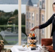 A hotel staff serves afternoon tea to guests by a large window overlooking a scenic view.