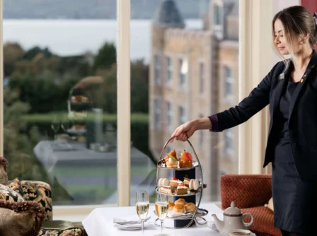 A hotel staff serves afternoon tea to guests by a large window overlooking a scenic view.