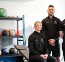 Three people in fitness gear smiling in a well-equipped gym at Hastings Hotels.