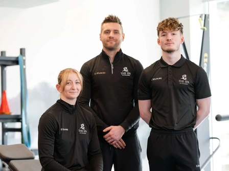 Three people in fitness gear smiling in a well-equipped gym at Hastings Hotels.