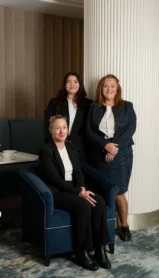 Three smiling women in a hotel lounge, dressed in formal attire, posing warmly.