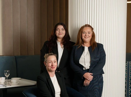 Three smiling women in a hotel lounge, dressed in formal attire, posing warmly.