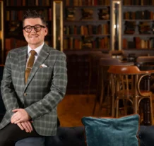 Stylish man in a plaid suit stands smiling in an elegant hotel lounge with bookshelves.
