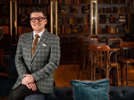 Stylish man in a plaid suit stands smiling in an elegant hotel lounge with bookshelves.
