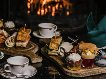 A cosy afternoon tea setup with sandwiches and pastries by a warm fireplace at a hotel.