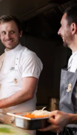 Two smiling chefs preparing dishes in a warmly lit, inviting kitchen.