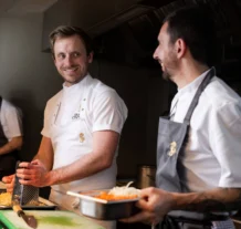 Chefs in a hotel kitchen preparing dishes, smiling and engaging warmly.