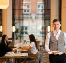 Waiter serves a couple in elegant hotel restaurant, large windows in the background.