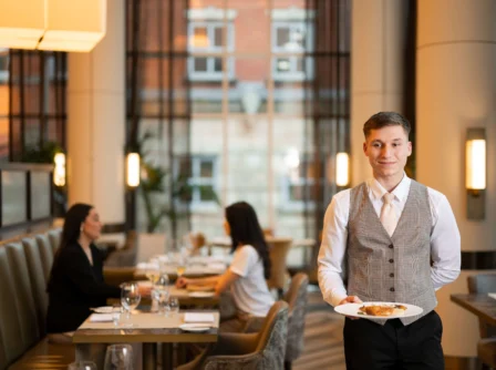 Waiter serves a couple in elegant hotel restaurant, large windows in the background.