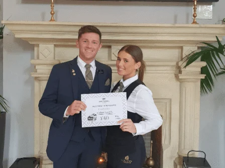 Smiling hotel staff in elegant uniforms holding a congratulatory certificate near a cosy fireplace.