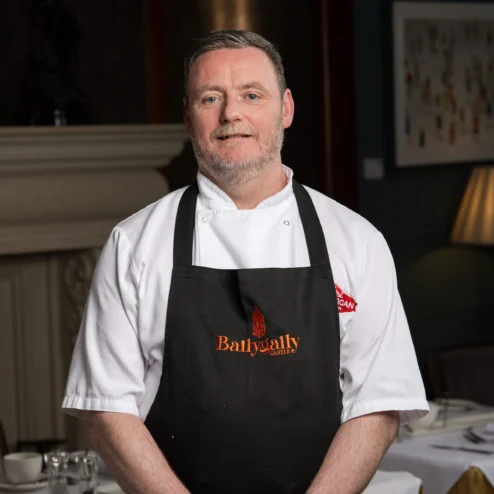 Chef in Ballygally apron stands in elegant dining room.