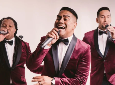 Three men in red velvet tuxedos singing joyfully with microphones.