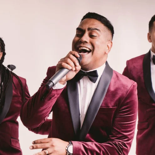 Three men in red velvet tuxedos singing joyfully with microphones.