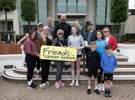 A joyful group stands outside Stormont Hotel, holding a 'Friends of the Cancer Centre' sign.