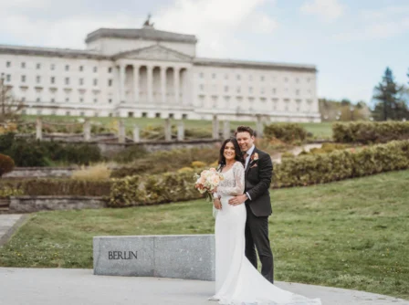 A joyful couple embraces in front of a grand building, holding a bouquet, surrounded by lush greenery.