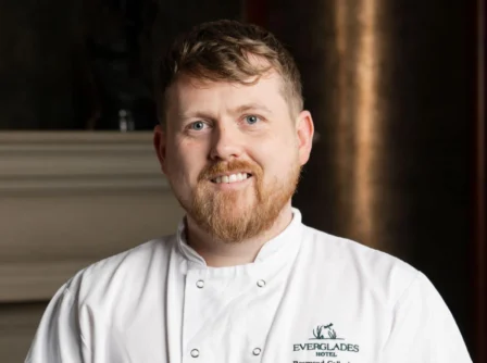 Chef in a white uniform warmly smiles in Everglades Hotel, fireplace in the background.