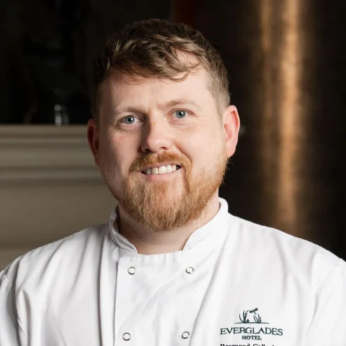 Chef in a white uniform warmly smiles in Everglades Hotel, fireplace in the background.