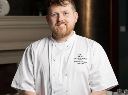Smiling chef in a hotel dining room, dressed in a white uniform, standing in front of a fireplace.