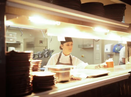 Chef handing meal to staff in warm, bustling hotel kitchen.