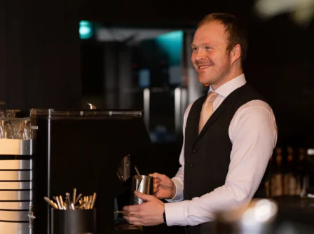 Smiling bartender at Hastings Hotels preparing a warm drink in a cosy bar setting.