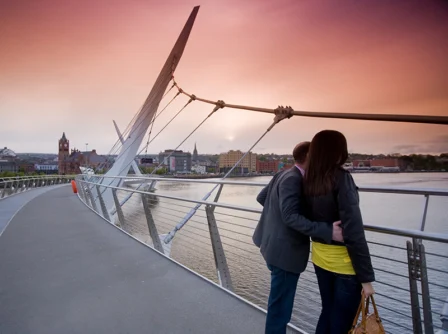 A couple embracing on a modern bridge at sunset, overlooking a scenic cityscape and river.