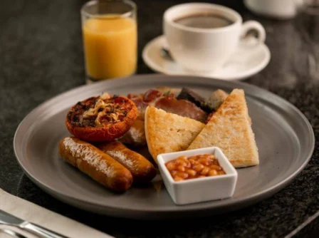 A full English breakfast with sausages, baked beans, grilled tomato, and hash browns, served with coffee and orange juice on a dark table.