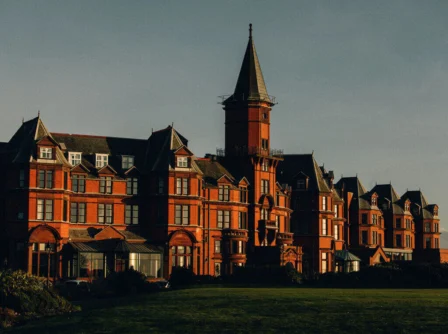 Elegant historic hotel with turret, surrounded by lush green gardens, under a clear sky.