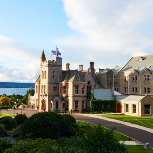 Elegant hotel by a sparkling sea with lush gardens and a vibrant blue sky.