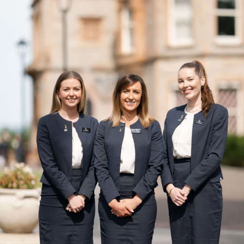 Three smiling hotel staff in navy uniforms outside an elegant brick building.