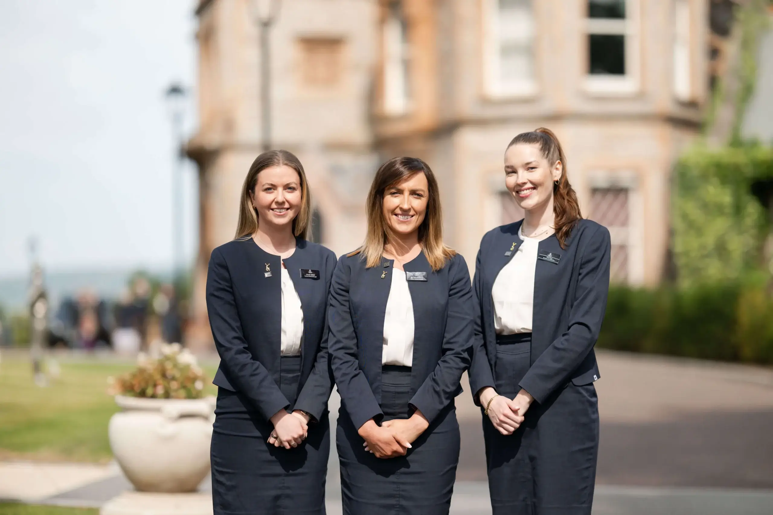 Three smiling hotel staff in navy uniforms outside an elegant brick building.