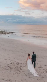 A couple walking on a serene beach at sunset, surrounded by calm sea and colourful skies.