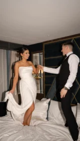 A couple in formal attire joyfully dance on a hotel bed in an elegant room.
