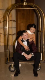 A couple in formal attire playfully sit on a luggage cart in a hotel lobby.