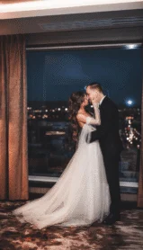 A couple in wedding attire embrace by a large window in a hotel room with city night views.