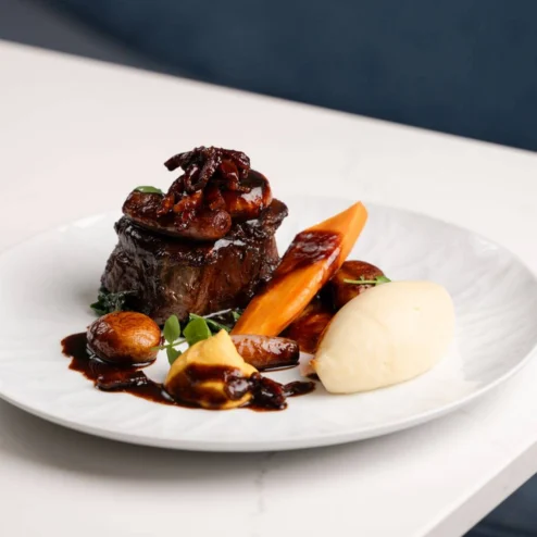 Succulent braised beef with glazed carrots, baby onions and creamy mash on a white plate in a hotel restaurant (no visible text)